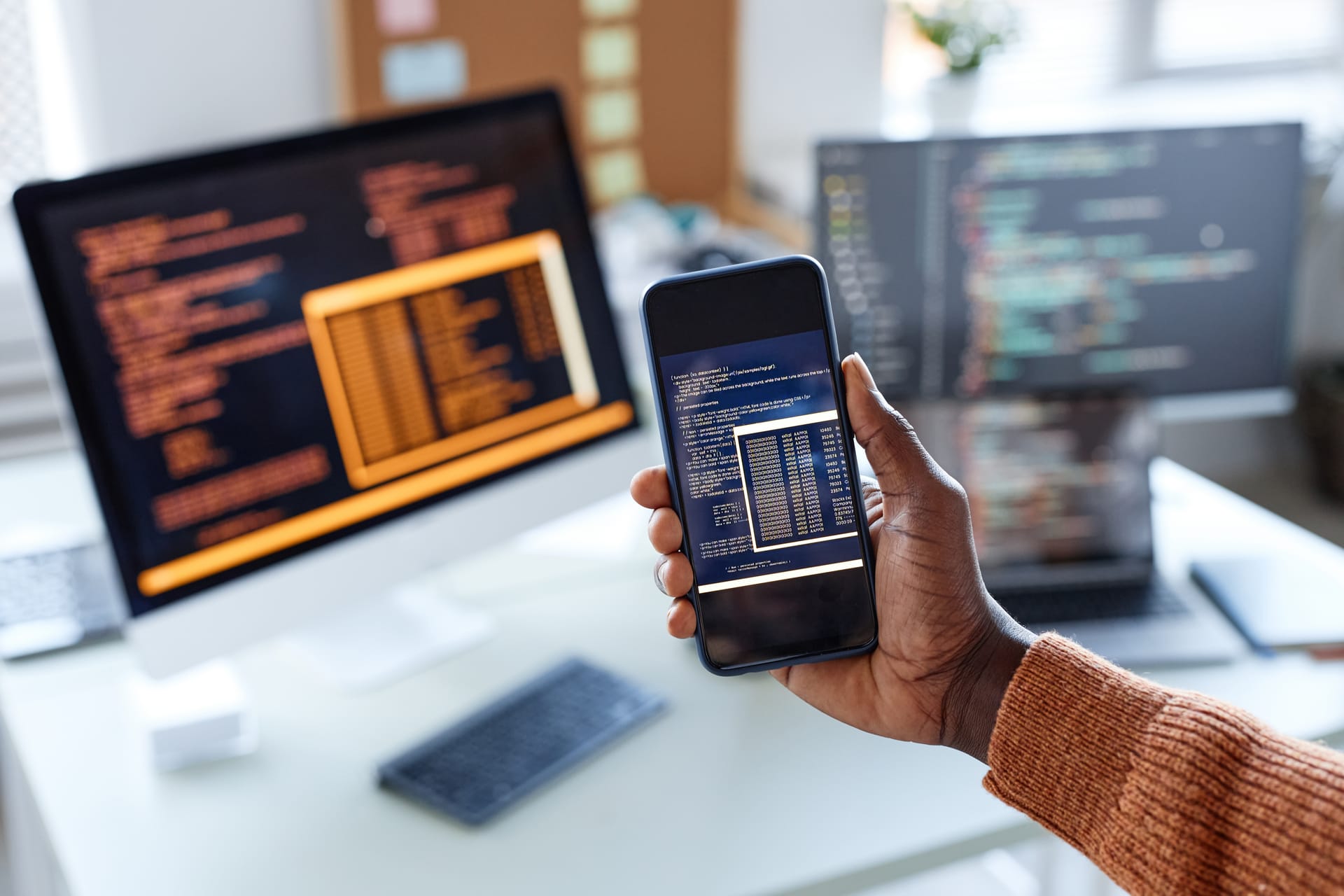 Developer holding a smartphone showing code beside monitors